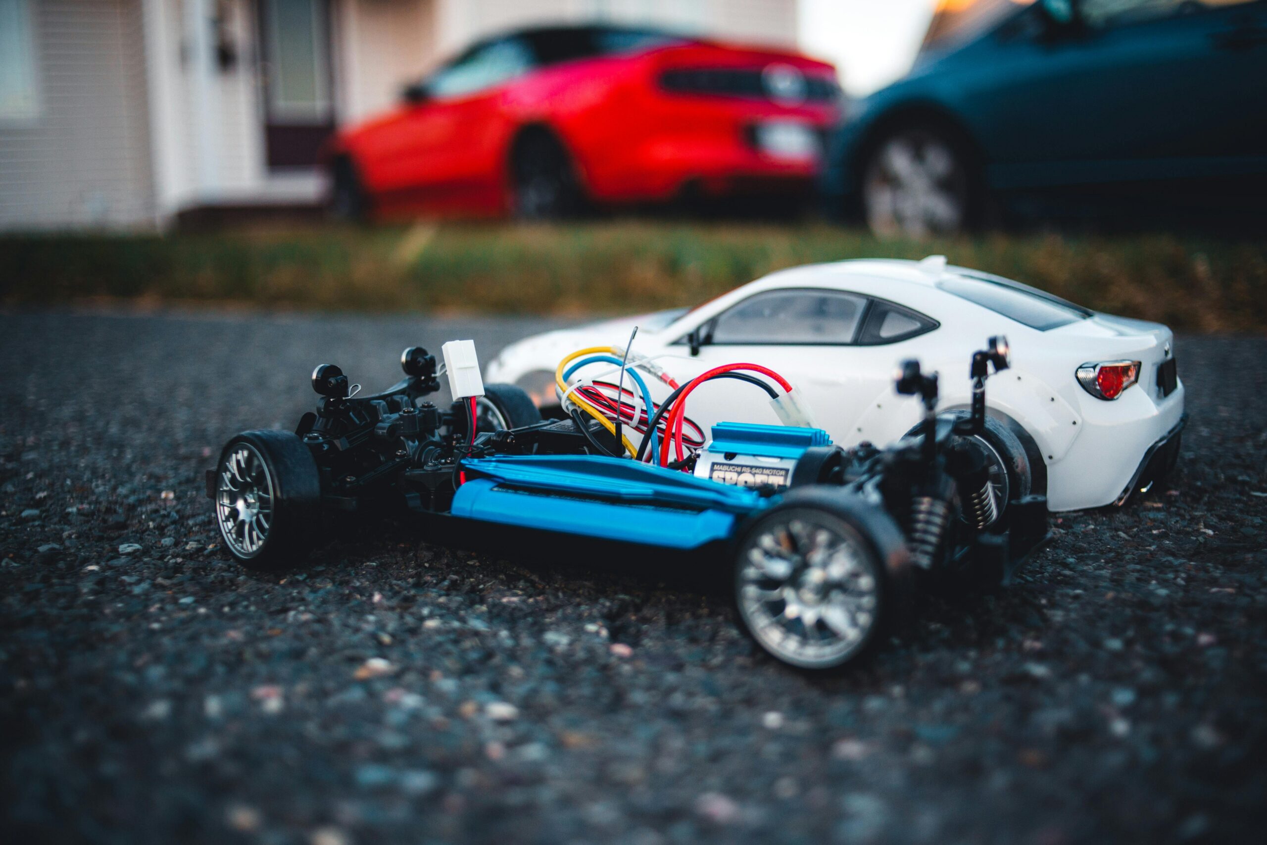 Close-up of a remote control car chassis with colorful wires on asphalt, with blurred cars in background.