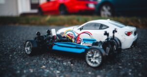 Close-up of a remote control car chassis with colorful wires on asphalt, with blurred cars in background.