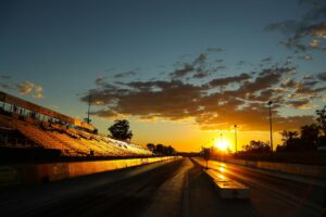 A stunning sunset illuminates an empty racetrack with bleachers, highlighting the sky and track lines.